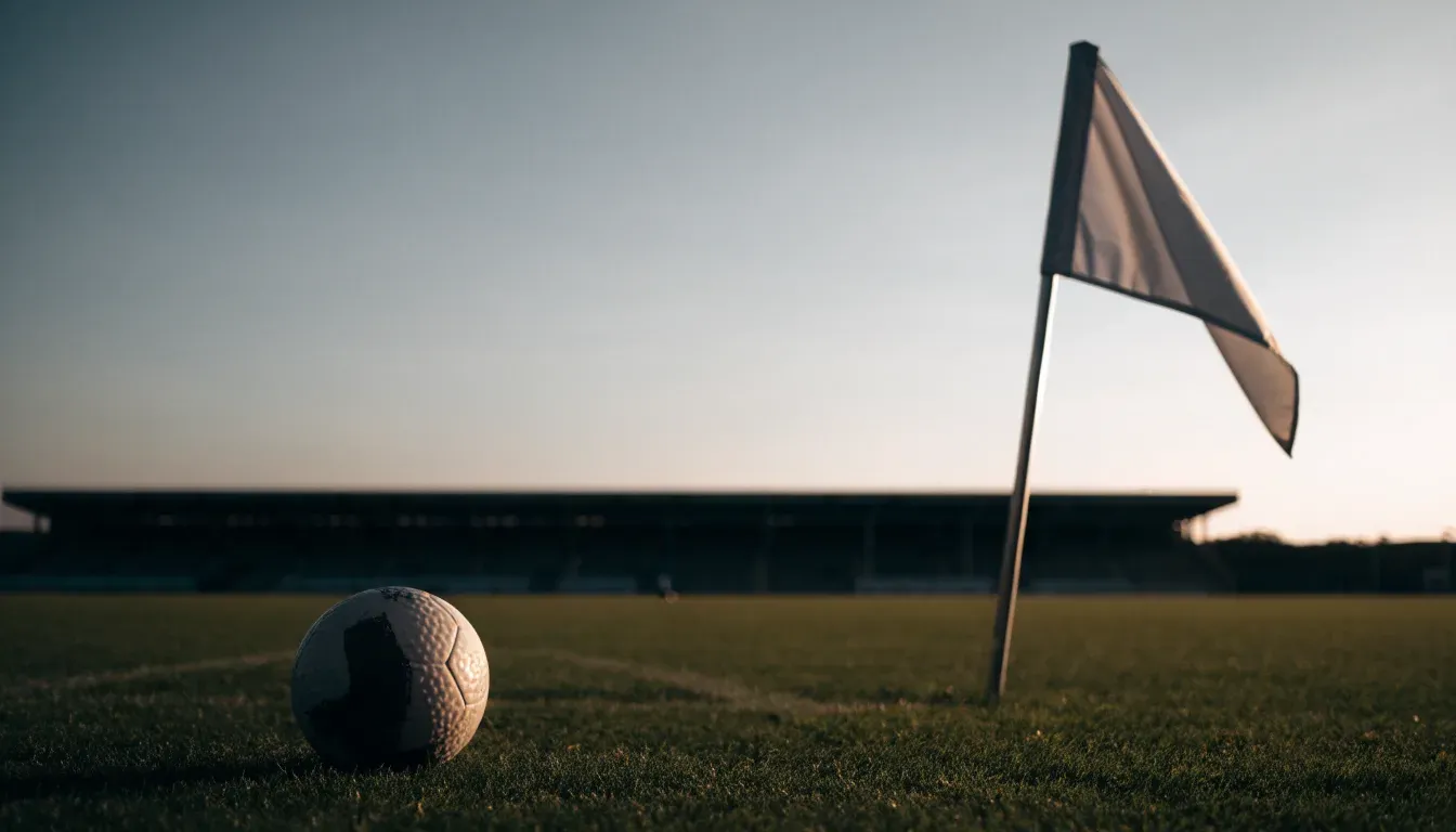 Ein Fußball liegt auf dem Rasen neben der Eckfahne eines leeren Platzes, dahinter eine Tribüne im Abendlicht.