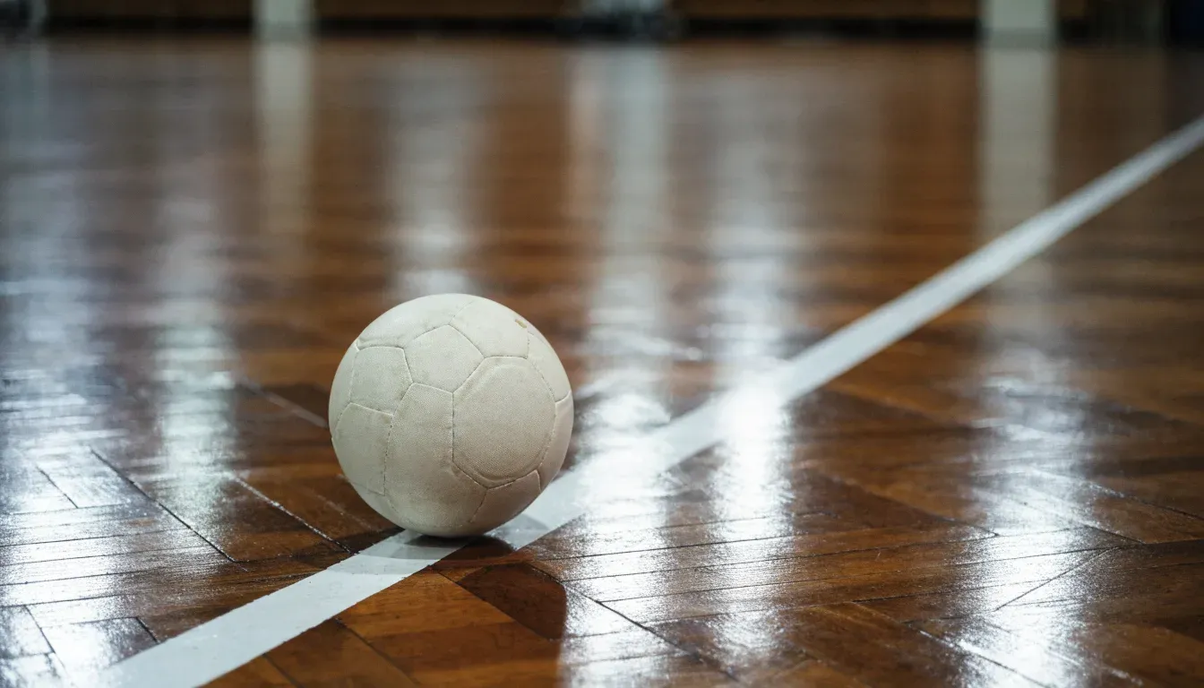 Ein Handball liegt auf dem Parkett einer Halle neben einer Spielfeldlinie, Licht fällt von oben ein.