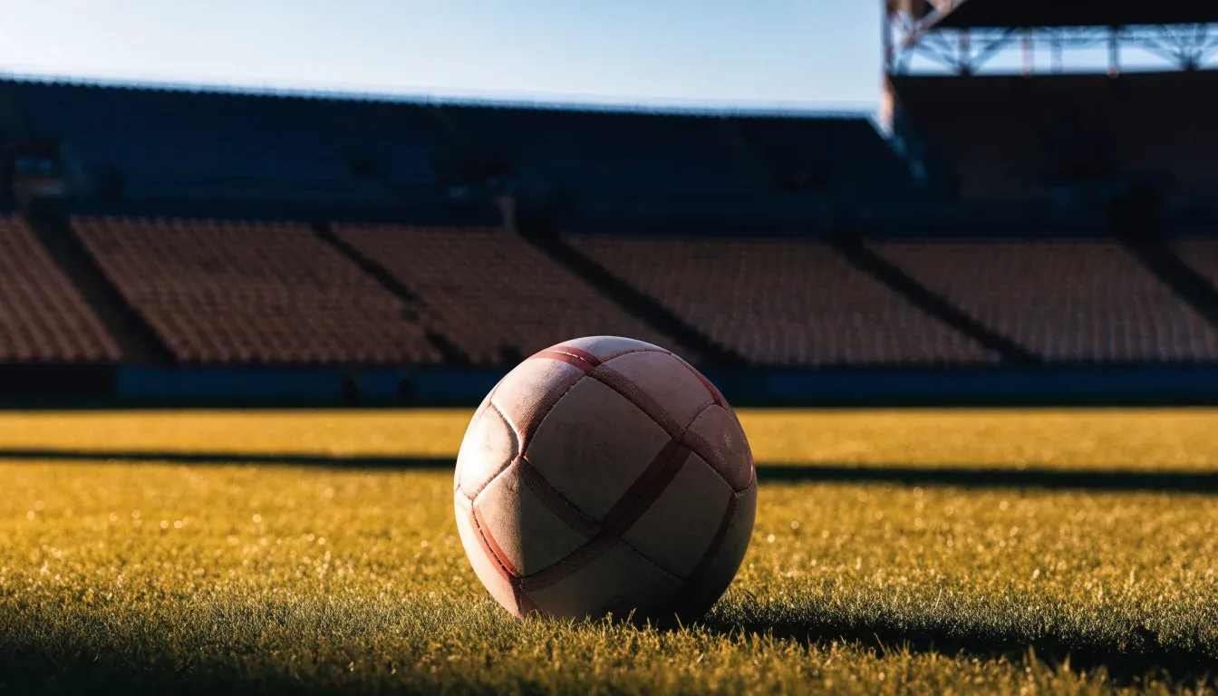 Ein Fußball liegt in der Mitte eines Rasens im Licht der tief stehenden Sonne, dahinter leere Sitzreihen.