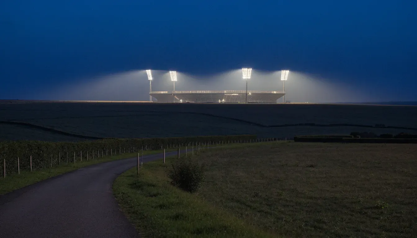 Ein Stadion unter Flutlicht aus der Ferne gesehen, davor eine dunkle Landschaft mit Feldern und einer Landstraße.
